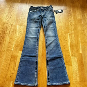 Ny True Religion Becca Bootcut Jeans Blå - Säljer ett par snygga True Religion Becca jeans i bootcut-modell. De är blå med coola slitningar och kontrastsömmar, har klassiska fem fickor och silverfärgade knappar. Midjan är midwaist och materialet är jeans med en mjuk känsla. Perfekt för dig som gillar retrovibbar och statement-jeans.