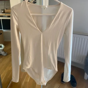 Vit långärmad v-ringad bodysuit - Säljer en stilren vit bodysuit från H&M med djup v-ringning och långa ärmar. Toppen är figurnära och tillverkad i mjuk bomull. Aldrig använd !!!