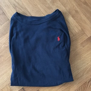 Marinblå långärmad tröja Polo Ralph Lauren - Snygg marinblå långärmad tröja från Polo Ralph Lauren med klassisk röd logobrodyr på bröstet. Tröjan är gjord i mjuk bomull och har en enkel, stilren design med rund hals. Perfekt för dig som gillar tidlös och clean stil.