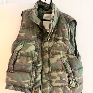 Camouflage dunväst från Ralph Lauren - Säljer en fet dunväst från Ralph Lauren Denim & Supply med klassiskt camouflage-mönster i gröna och bruna toner. Västen har hög krage, stora fickor med lock och både dragkedja och knappar framtill. Perfekt för lager-på-lager och streetwear-vibe.