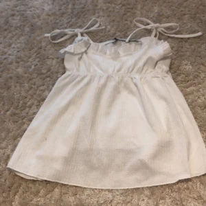 Vit linnetopp med knytband XS - Supersöt vit linnetopp från 157 i storlek XS. Toppen har smala axelband med knyt och en lätt rynkad volangkant upptill. Materialet är mjukt och har en diskret ribbad struktur som ger en chill vibe. Perfekt för varma dagar och enkel att matcha med jeans eller kjol.