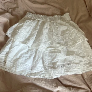 Vit volangkjol med spetsdetaljer - Superfin vit kortkjol med volanger och spetsdetaljer. Kjolen har resår i midjan och är gjord i ett lätt bomullstyg, perfekt för varma dagar. Söt och romantisk stil med broderade kanter och små bollar längst ner.