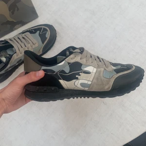 Valentino Rockrunner sneakers camo - Snygga Valentino Rockrunner sneakers med grå, beige och svarta camouflage-detaljer. Skorna har svarta och beige partier i mocka och skinn, samt klassisk snörning och svart sula. Skorna kommer med box, dustbag och extra snören. Har använts färgen har slitits av lite på toppen (se bild 5) annars är dom i väldigt bra skick, hör av dig om du har frågor. pris kan diskuteras