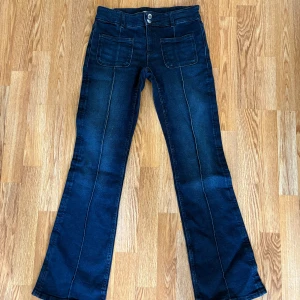 Mörkblå bootcut jeans med fickor - Snygga mörkblå jeans med bootcut passform och markerade sömmar längs benen. Byxorna har två stora framfickor och klassisk knappstängning. Perfekta för dig som gillar en retroinspirerad look.