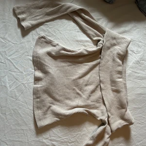 Beige ribbad omlottop från Gina Tricot - Supersnygg beige ribbad omlottop från Gina Tricot. Toppen har långa ärmar och knyts i midjan för en trendig look. Perfekt att styla med jeans eller kjol för en chill men ändå uppklädd vibe.