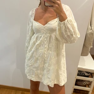 Vit blommig kortklänning med puffärm - Supersöt vit kortklänning med broderade blommor och puffiga långärmar. Klänningen har v-ringning, knappar framtill och en flowy passform som ger en romantisk vibe. Perfekt för varma dagar eller när du vill känna dig extra fin.