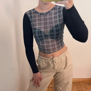 Rutig mesh topp från Zara - Säljer en croppad långärmad topp från Zara i mesh med rutigt mönster i grått och svart. Toppen har svarta ribbade ärmar och transparent rutigt tyg framtill och baktill. Perfekt för dig som gillar edgy och trendiga plagg.