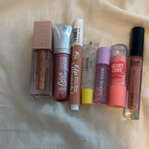 Mix av läppglans / läppoljor och lip balm  - Säljer ett kit med sju olika läppglans och läppoljor i olika färger och former. Inkluderar Maybelline Lifter Gloss i brons, W7 Lip Punch i nude, Essence Heart Core i rosa, samt grape soda och lemon lip oil. Flera gloss har shimmer eller metallic finish. Perfekt för dig som vill testa olika looks vissa är lite gammlare än de andra men super fina 