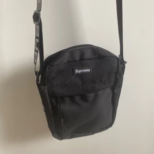Svart axelväska från Supreme - Supreme axelväska i svart med justerbart band där loggan är tydligt tryckt i vitt. Väskan har dragkedja upptill och en extra ficka framtill. Perfekt storlek för det viktigaste och en clean streetstyle-look.