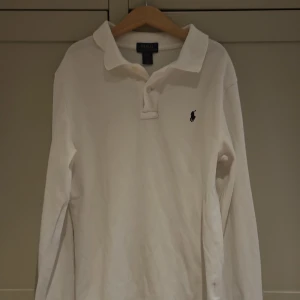 Vit pikétröja från Ralph Lauren - Klassisk vit långärmad pikétröja från Ralph Lauren med ikonisk broderad logga på bröstet. Tröjan har krage och knäppning framtill, perfekt för en clean och stilren look. Tillverkad i mjuk bomull som känns skön mot huden.