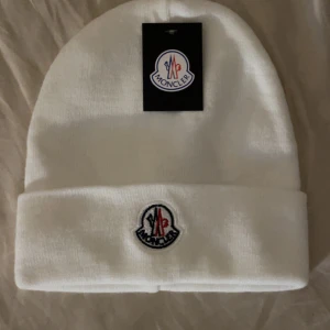 Vit Moncler mössa med broderad logga - Vit mössa från Moncler med klassisk uppvikt kant och broderad logotyp framtill. Stilren och enkel design som passar till det mesta. Perfekt accessoar för kyliga dagar. FINNS ÄVEN I SVART