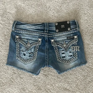 Miss Me blå jeansshorts med strass - Snygga blå jeansshorts från Miss Me med slitna detaljer, råa kanter och blingade bakfickor med strass och broderier. Shortsen har låg midja, dekorativa sömmar och coola nitar. Perfekta för dig som vill sticka ut med en edgy och glam stil. Midjemått: 72cm 💞