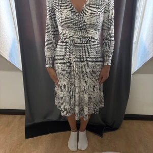 Svartvit omlottklänning med knyt - Snygg svartvit omlottklänning med grafiskt rutigt mönster. Klänningen har v-ringning, långa ärmar och ett knytband i midjan som framhäver figuren. Mjuk och följsam passform, perfekt för dig som gillar stilrena och trendiga plagg.