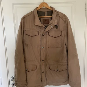 Beige jacka från Massimo Dutti - Stilren beige jacka från Massimo Dutti med fyra fickor framtill och snygga detaljer vid axlar och krage. Jackan har knappar och dragkedja, samt hög krage med spänne. Perfekt för dig som gillar klassisk och clean look. Nypris 1799
