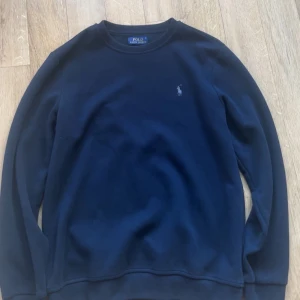Mörkblå sweatshirt från Polo Ralph Lauren - En stilren mörkblå sweatshirt från Polo Ralph Lauren med klassisk rund hals och broderad logga på bröstet. Tröjan har långa ärmar och är tillverkad i mjuk bomull, perfekt för en chill och snygg look.
