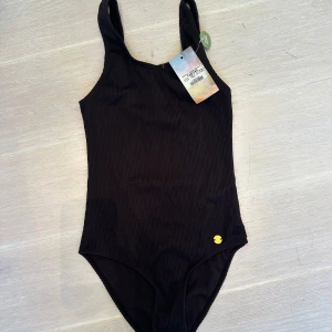 Svart ribbad oanvänd baddräkt från KappAhl - Svart baddräkt med ribbad struktur och klassisk skärning från KappAhl med liten gulddetalj i hörnet. Helt oanvänd+prislapp!