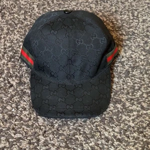 Svart Gucci keps med GG-mönster - Snygg svart keps från Gucci med klassiskt GG-monogram över hela kepsen och ikoniska gröna och röda ränder på sidorna. Justerbar rem baktill för perfekt passform. Tillverkad i Italien och i ett slitstarkt bomullsmaterial.