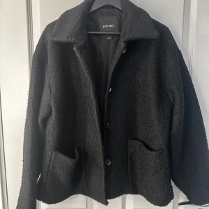 Svart jacka från Monki - Stilren svart jacka från Monki med grov textur. Klassisk krage, stora fickor framtill och knäppning med tre knappar. Perfekt för lager-på-lager och ger en chill men ändå snygg vibe till din outfit.