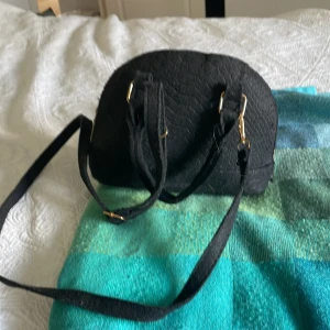 Svart axelväska i filtmaterial - Säljer en svart axelväska med rundad form och guldfärgade metalldetaljer. Väskan har både handtag och lång axelrem samt dragkedja upptill. Ytan är i filtliknande material med diskret struktur. Perfekt storlek för det viktigaste du vill ha med dig. Man kan ta av axelbandet!