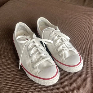 Vita Converse All Star sneakers - Klassiska vita Converse All Star sneakers i canvas med lågt skaft. Skorna har vit platt sula med röd och blå rand, rund tå och snörning framtill. Perfekta för en avslappnad och cool stil.