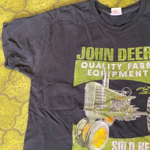 John Deere svart t-shirt med tryck - Svart t-shirt med stort vintageinspirerat John Deere-tryck i grönt och vitt framtill. Motivet visar en klassisk traktor och texten 'Quality Farm Equipment'. Rund halsringning och korta ärmar. Size L 