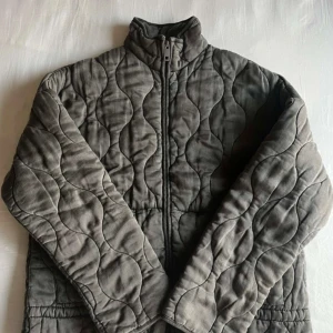 Grå quiltad jacka med dragkedja - Quiltad jacka i grått med vågigt mönster och hög krage. Jackan har hel dragkedja framtill och är tillverkad i ett mjukt bomullstyg. Perfekt för dig som gillar en clean och chill look med lite edge.