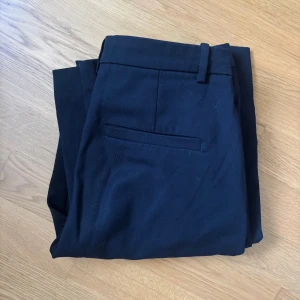 Svarta kostymbyxor från LINDEX - Snygga svarta kostymbyxor från LINDEX med klassisk passform och raka ben. Byxorna har hällor för bälte, sidfickor och en bakficka. Perfekta för en clean och stilren look. Materialet känns mjukt och följsamt.