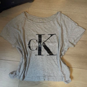 Grå Calvin Klein Jeans t-shirt - Snygg grå t-shirt från Calvin Klein Jeans med stort svart CK-tryck framtill. Modellen är croppad och har korta ärmar och rund halsringning. Tillverkad i mjuk bomull, perfekt för en avslappnad och trendig look. Litet hål på tröjan (se sista bilden). 