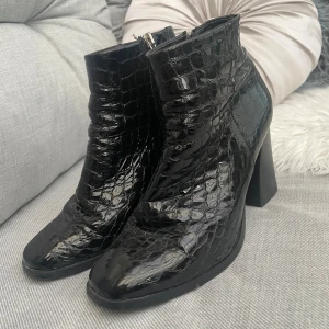 Svarta boots med krokopräglat mönster Mingo - Snygga svarta boots från Mingo med krokopräglat mönster i glansigt skinn. Skorna har en rejäl blockklack, rund tå och dragkedja på insidan. Perfekta för dig som vill sticka ut med en trendig och edgy look. BARA POSTNORD