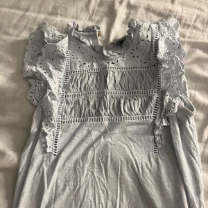 Vit blus med volang från Holly & Whyte - Supersöt vit blus från Holly & Whyte med broderade detaljer och volang vid axlarna. Blusen har rundad hals med liten volangkant, knapp i nacken och är ärmlös. Perfekt under alla säsonger!