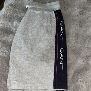 Grå GANT shorts med logga och snörning - Snygga grå shorts från GANT med breda mörkblå sidor och röd kantlinje. GANT-loggan syns tydligt längs båda sidorna. Mjuk bomullskänsla, elastisk midja med snörning och två fickor framtill. Perfekta för chill dagar eller träning.