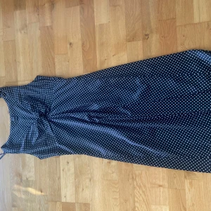 Svart prickig långklänning Esprit - Snygg svart långklänning från Esprit med vita prickar. Klänningen är ärmlös och har en klassisk, feminin siluett med lätt markerad midja. Materialet är glansigt och känns lyxigt mot huden. Perfekt för dig som gillar retrovibbar och tidlös stil.