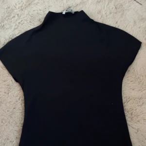 Svart polotopp från H&M XS - En stilren svart halvkrage topp från H&M i storlek XS. Toppen har korta ärmar och hög hals, vilket ger en clean och modern look. Perfekt att matcha med jeans eller kjol för en snygg outfit.