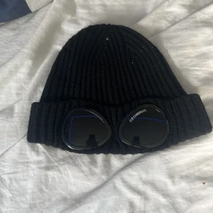 Svart C.P. Company goggle-mössa -  Pris kan diskuteras  Ribbstickad svart mössa från C.P. Company med ikoniska goggle-linser framtill. Mössan är tillverkad i mjukt material och har en bred uppvikt kant. Perfekt för dig som vill sticka ut med streetwear-vibe och klassisk design.