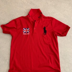Röd Polo Ralph Lauren England t-shirt - Röd t-shirt från Polo Ralph Lauren med broderad engelsk flagga och texten ENGLAND på bröstet. Stor svart polospelare-logga, nummer 5 på ärmen och tryck på kragen. Klassisk pikékrage och korta ärmar. Perfekt för dig som gillar sportig och stilren look.