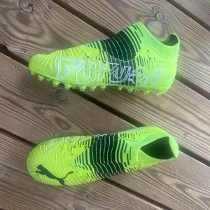 Neongula Puma Future fotbollsskor - Säljer ett par riktigt snygga Puma Future fotbollsskor i neongult med svarta detaljer och vit text. Skorna har en strumpliknande passform utan snörning och är designade för gräsplan. Perfekta för dig som vill sticka ut på planen!