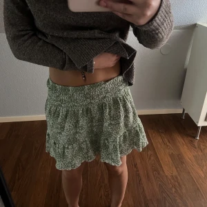 Grön blommig volangkjol - Supersöt grön kortkjol med vitt blommönster och volangkant. Kjolen har smock i midjan för en bekväm passform och är luftig och lätt, perfekt för varma dagar. Passar dig som gillar en avslappnad och trendig stil.