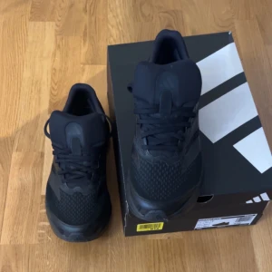 Svarta Adidas sneakers med snörning - Snygga helsvarta Adidas sneakers med klassiska tre ränder på sidan. Skorna har snörning och en sportig, modern design med rund tå och platt sula. Perfekta för dig som gillar stilrena och bekväma skor till vardags eller träning.