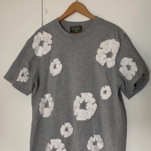 Grå t-shirt med vita blommor Denim Tears - Grå t-shirt från Denim Tears med stora vita blommiga tryck över hela plagget. Klassisk passform med rund hals och korta ärmar. Tillverkad i 100% bomull för en skön känsla. Perfekt för dig som vill sticka ut med en unik och trendig look.