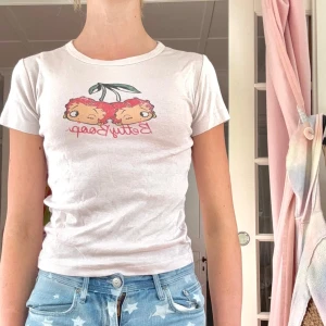 Ljus rosa Betty Boop t-shirt Fleischer XS - 2000-T-shirt från Fleischer Studios med Betty Boop-tryck i form av två körsbär och röd text. Modellen är figurnära med korta ärmar och rund halsringning. Perfekt för dig som gillar retro vibes och coola prints.
