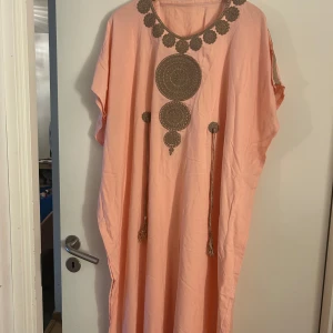 Ljusrosa långklänning med broderi - Lång, loose-fit klänning i ljusrosa bomull med stora bruna broderade cirklar och dekorativa tofsar framtill. Rund halsringning med broderade detaljer längs kanten. Perfekt för dig som gillar boho-vibe och unika detaljer.marockans 