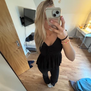 Svart blus med snörning från 157 - Supersöt svart blus från 157 med v-ringning och snörning framtill. Toppen har en lätt volang nertill och är ärmlös, perfekt för att styla med jeans eller kjol. Materialet är mjukt och har en lätt struktur som ger en snygg look.