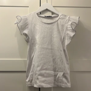 Vit topp med volangärmar - Supersöt vit topp med volangärmar och kontrastsömmar i svart längs kanterna. T-shirten har rund halsringning och är tillverkad i ett mjukt, ribbat material. Är i storlek S men passar även Xs💛