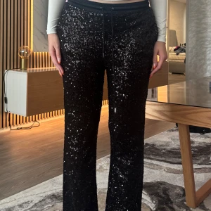 Svarta paljettbyxor med snörning - Glittriga svarta byxor täckta med paljetter och elastisk midja med snörning. Byxorna har en rak passform och ger en riktigt festlig vibe. Perfekta för dig som vill sticka ut och glänsa lite extra. Bra för xs-s
