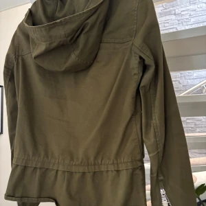 Olivgrön jacka med huva - Snygg olivgrön jacka med huva och två stora fickor framtill med dragkedjor. Jackan har en lätt rynkad nederkant och stängs med både dragkedja och tryckknappar. Perfekt för dig som gillar en avslappnad men ändå stilren look.