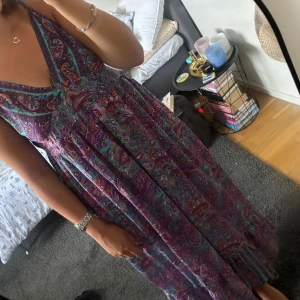 Lång Ibiza klänning i lila - Säljer en lång, flowy klänning med paisleyinspirerat mönster i lila, turkos och orange. Djup v-ringning, smala justerbara axelband och resår i ryggen, vilket gör att den passar XS-L. Funkar som kjol och klänning.