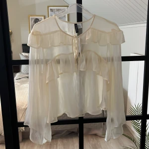 Vit volangblus från H&M Divided - Superfin vit blus från H&M Divided i transparent, lätt material. Blusen har långa ärmar, volangdetaljer över axlar och bröst samt knytning framtill vid halsen. Oanvänd, fortfarande prislapp kvar. 