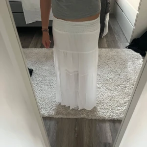 Vit långkjol från H&M med smock - Supersöt vit långkjol från H&M med bred smock i midjan. Kjolen har flera volanger. Och har en Välldigt liten obehandlad fläck på sidan. I storlek 158