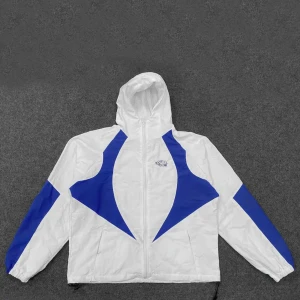 Vit och blå vindjacka med huva - Snygg vit vindjacka med blåa detaljer och huva. Jackan har dragkedja framtill, elastiska muddar och coolt grafiskt mönster på ärmar och bröst. Perfekt för dig som gillar sportig och retro stil. Lätt och smidig att bära, passar till streetwear och chill dagar.