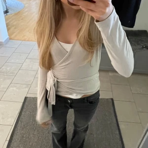 Ljus beige omlottopp med knyt - Supersnygg ljus beige omlottopp med långa ärmar och knyt i sidan. Toppens v-ringning och figurnära passform ger en clean och modern look. Perfekt att styla med jeans eller kjol för en trendig outfit.